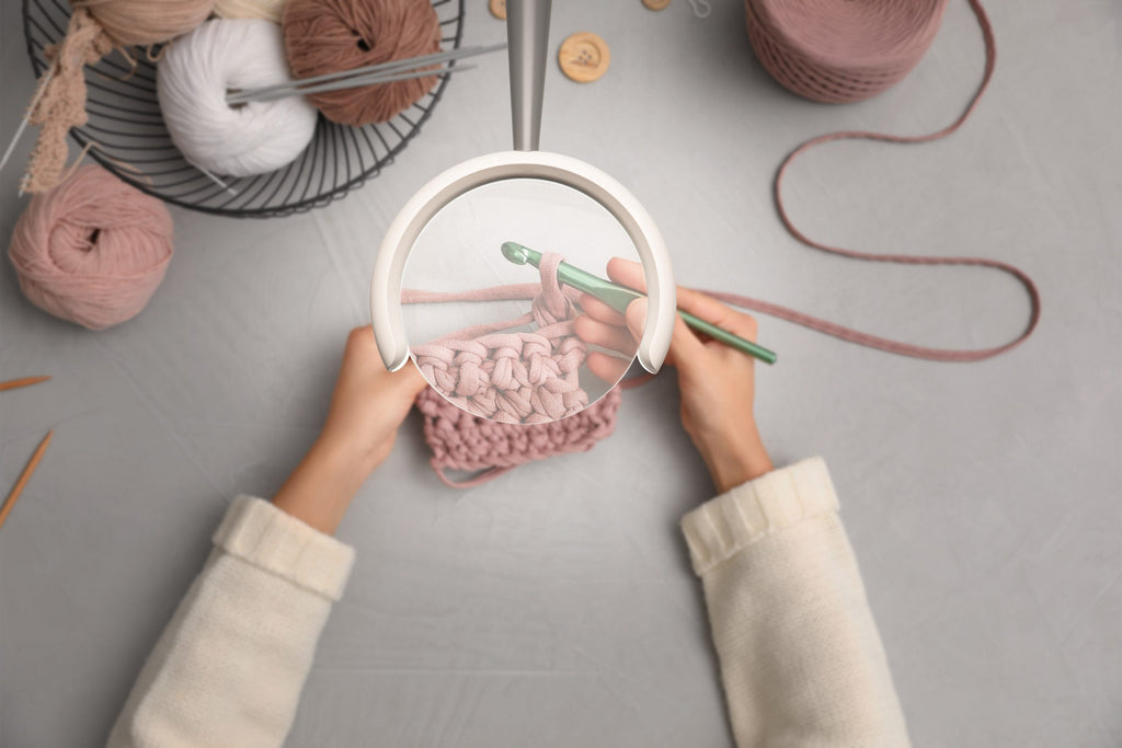 Top view of woman knitting with pink yarn on a gray surface. We see the portion under the lamp is magnified. 