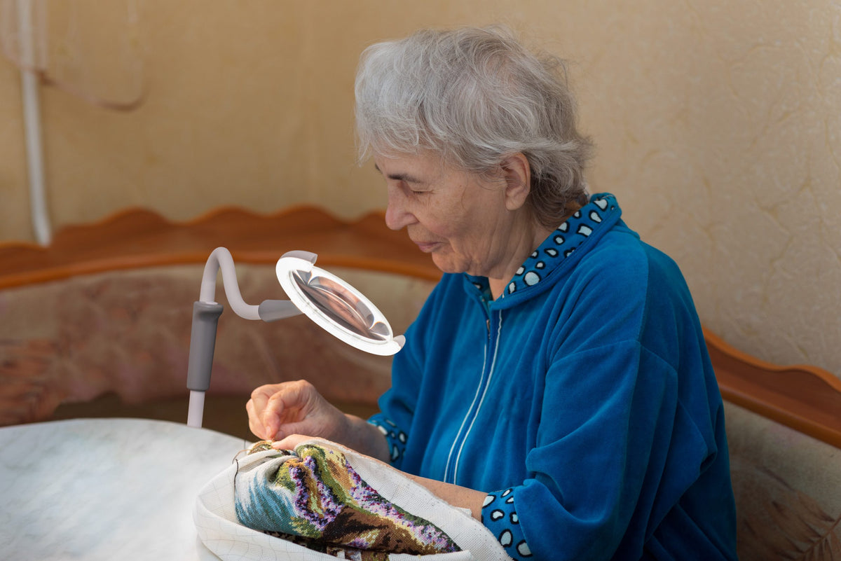 Woman in blue jacket using a magnifying lamp to work on a craft project.