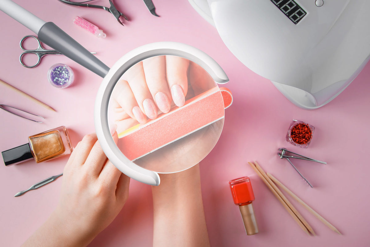 A top-down view of a manicure setup on a pink background, featuring a hand under a magnifying glass using an orange nail file, with nail polish bottles, tools, and a white nail dryer arranged around it.
