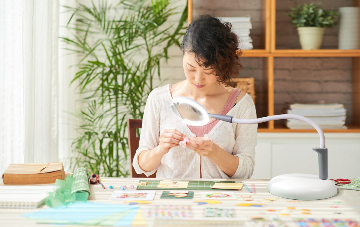 Woman sitting at a table with craft materials, using the magnifying lamp to work.