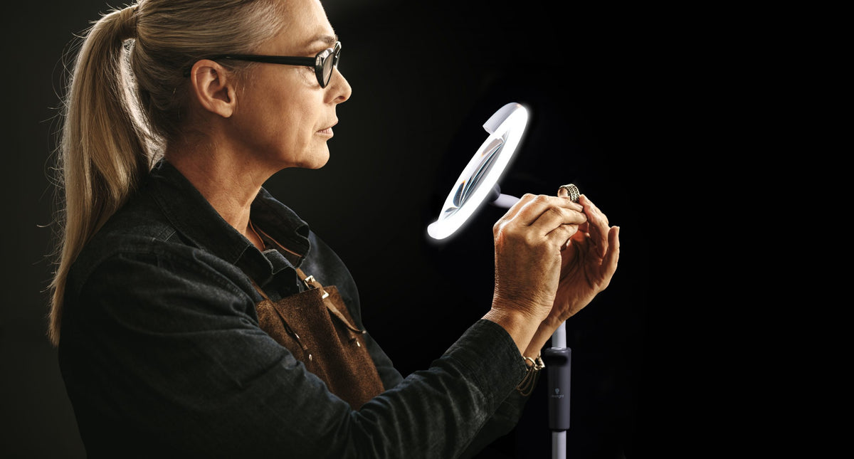 Woman wearing glasses holding a small object under a magnifying lamp on a black background