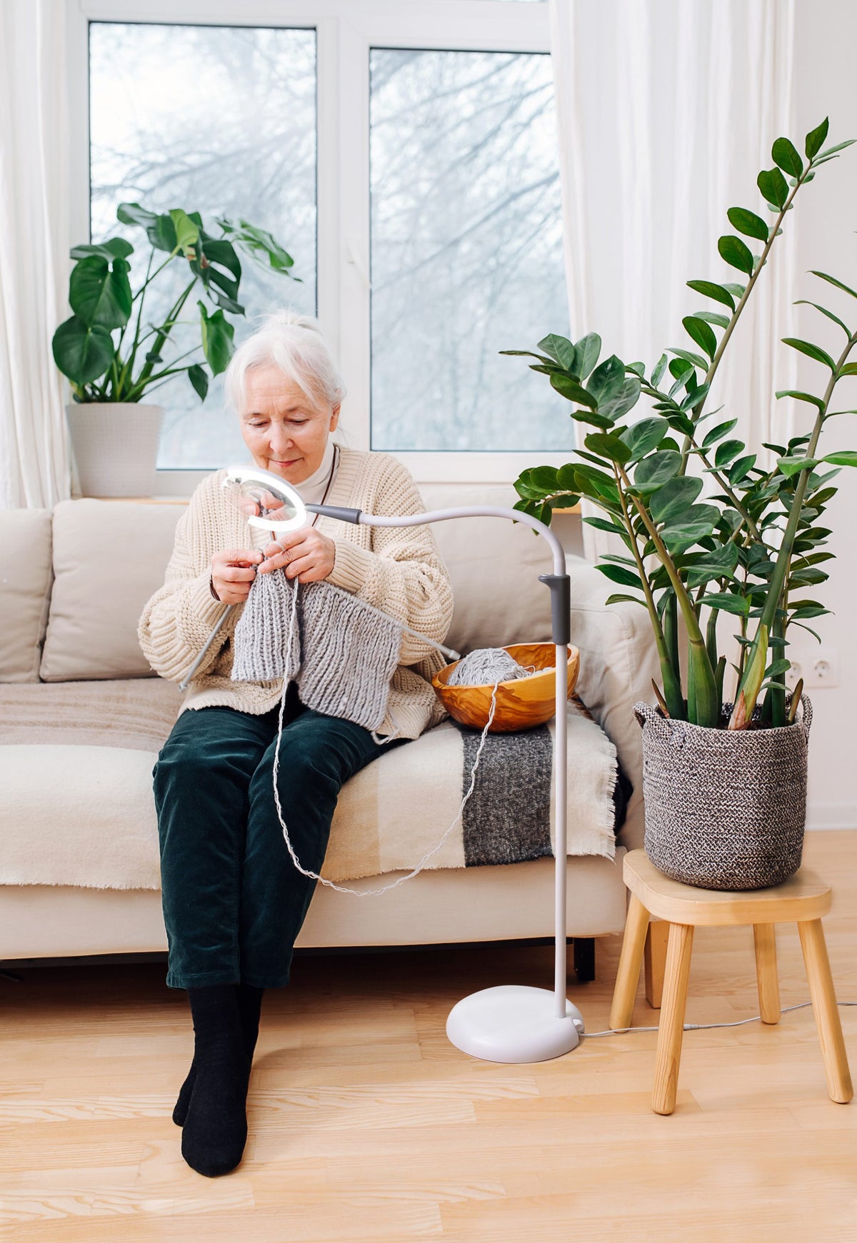 Senior woman knitting on a couch with magnifier lamp close to her hands.