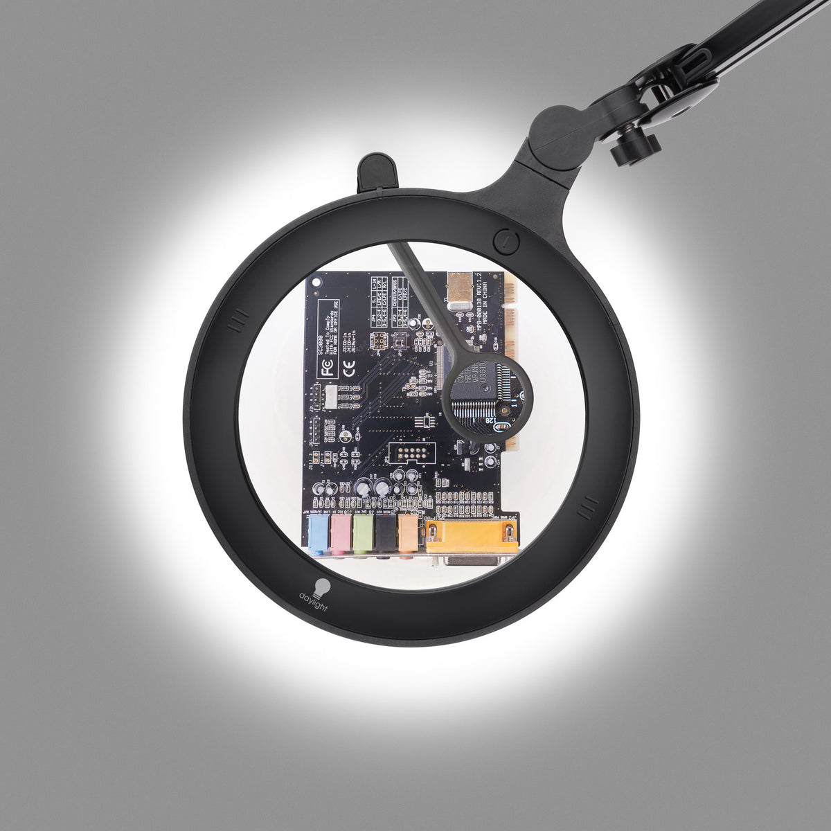 A black magnifying ring light with an integrated LED light and adjustable arm, shown magnifying a circuit board underneath.