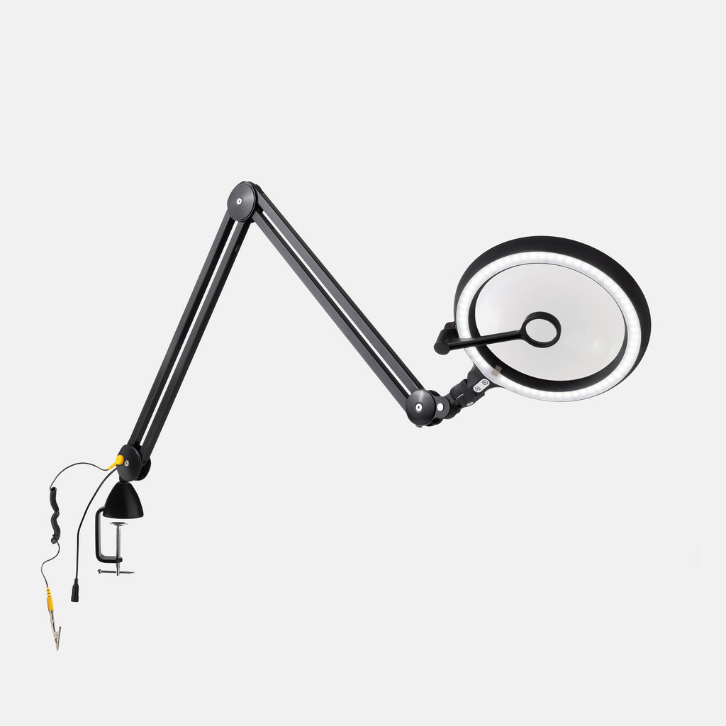 A black adjustable desktop lamp with a circular magnifying lens, flexible articulated arm, and clamp base, shown on a plain light gray background.