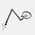 A black adjustable desktop lamp with a circular magnifying lens, flexible articulated arm, and clamp base, shown on a plain light gray background.