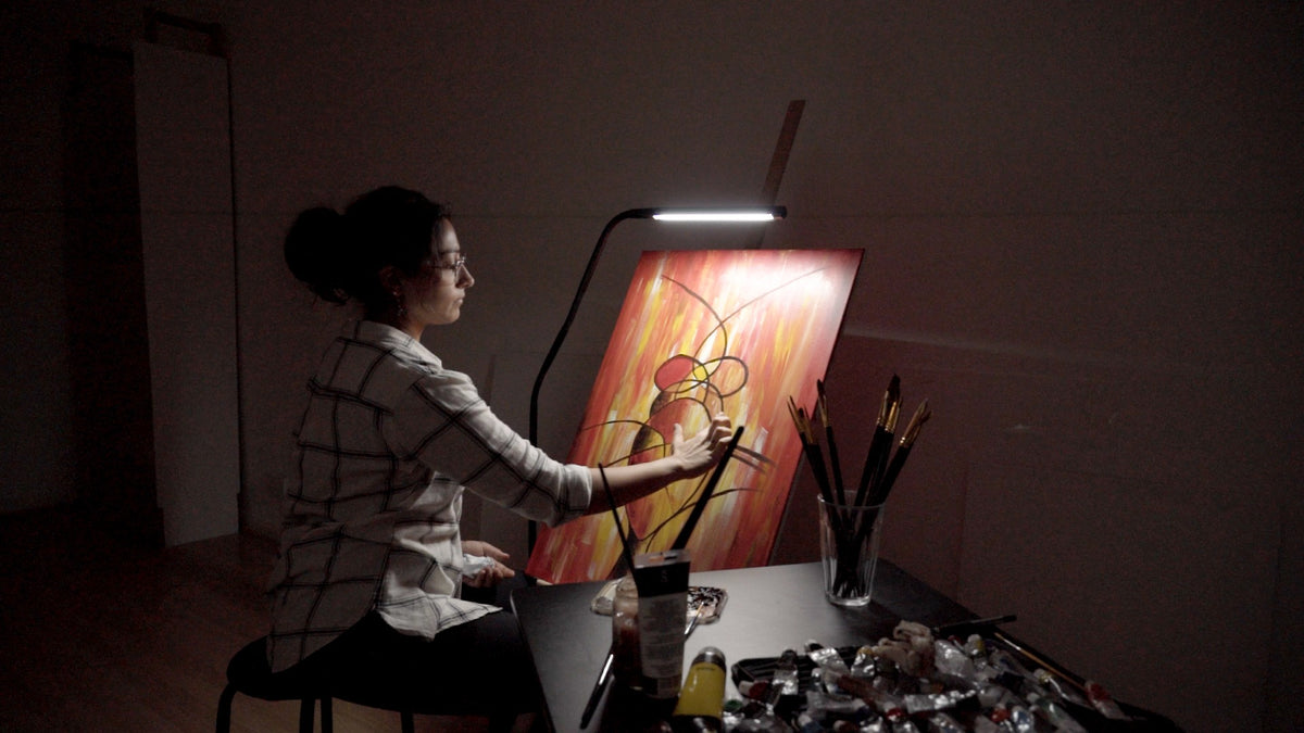 A woman wearing glasses painting at a table under a bright desk lamp, with a colorful abstract painting on the canvas and paintbrushes in a glass vase.