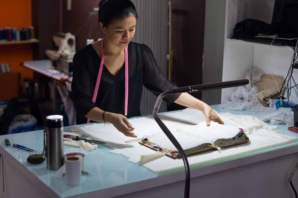 Woman working at a table with sewing materials and tools. Light is bent over pattern book. 
