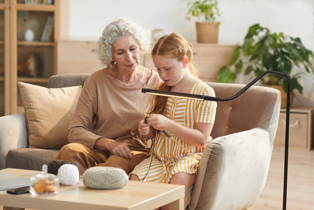 Woman and young girl sitting on a couch in a living room, with the woman teaching the girl to knit. Light is bent close to her hands.