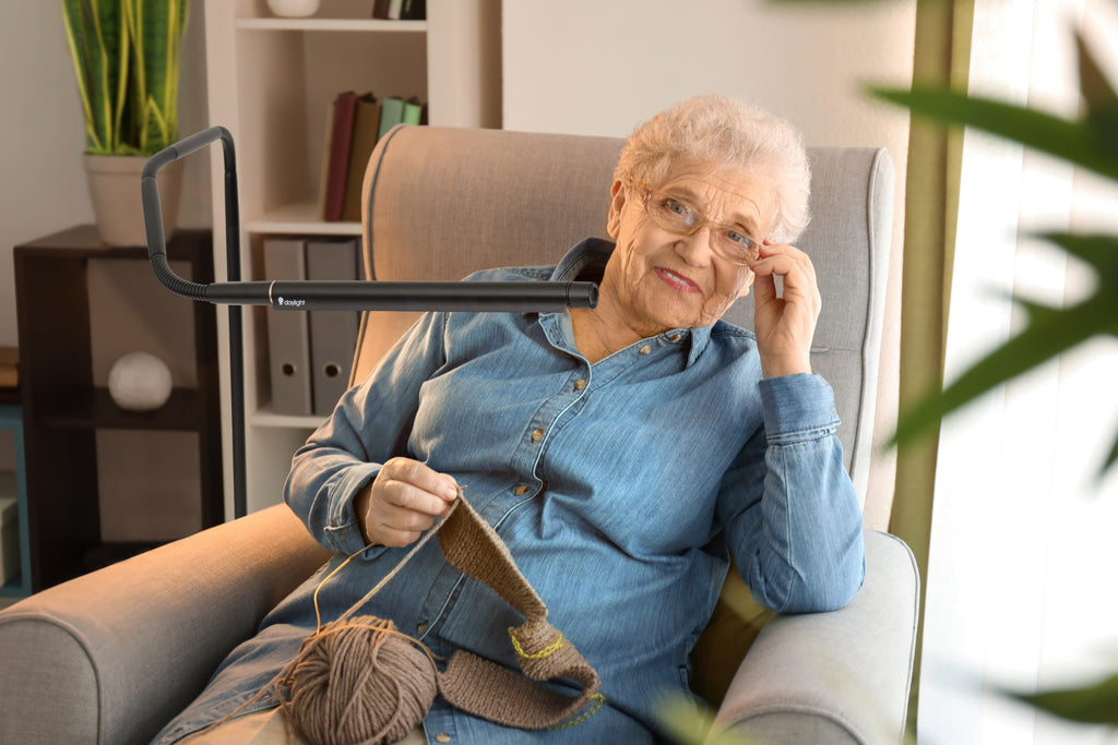 Senior woman sitting in a chair with knitting materials, wearing glasses. Light is bent close to the knitting. 