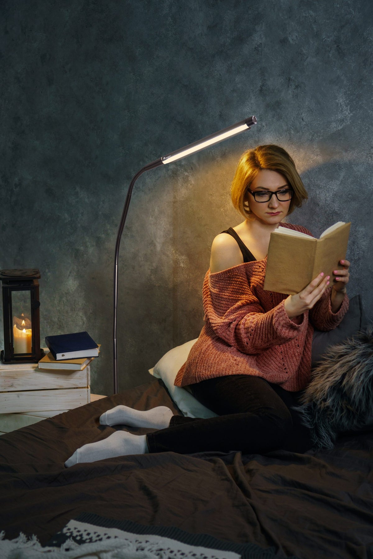 A woman with short blonde hair and glasses sits on a bed reading a book, illuminated by a floor lamp bent over the book. 