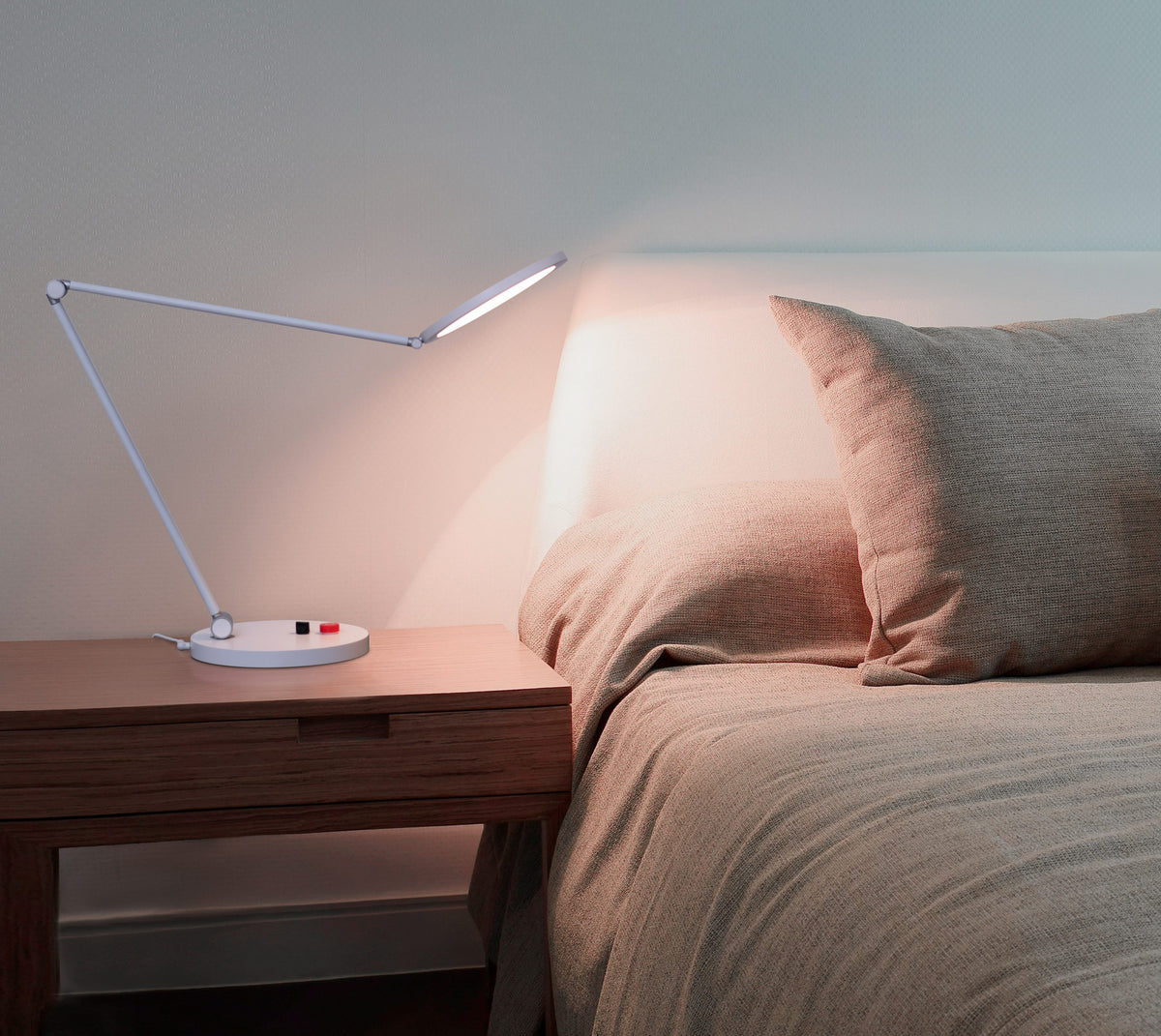 Foldable arm desk lamp on a nightstand next to a bed with beige bedding.
