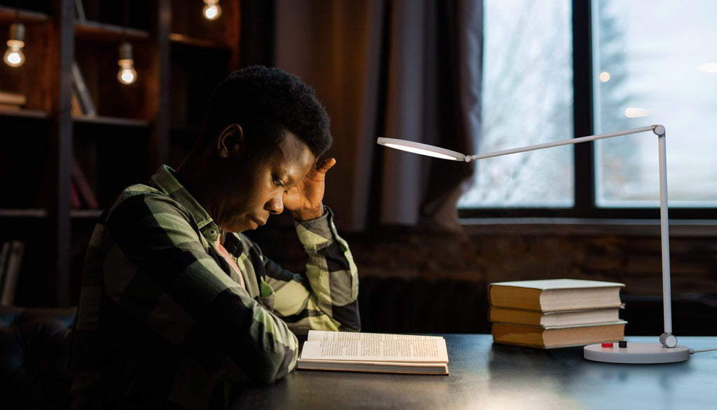 A young man in a green camouflage shirt sits at a desk reading an open book under a white adjustable desk lamp, with a stack of books nearby and a window in the background.