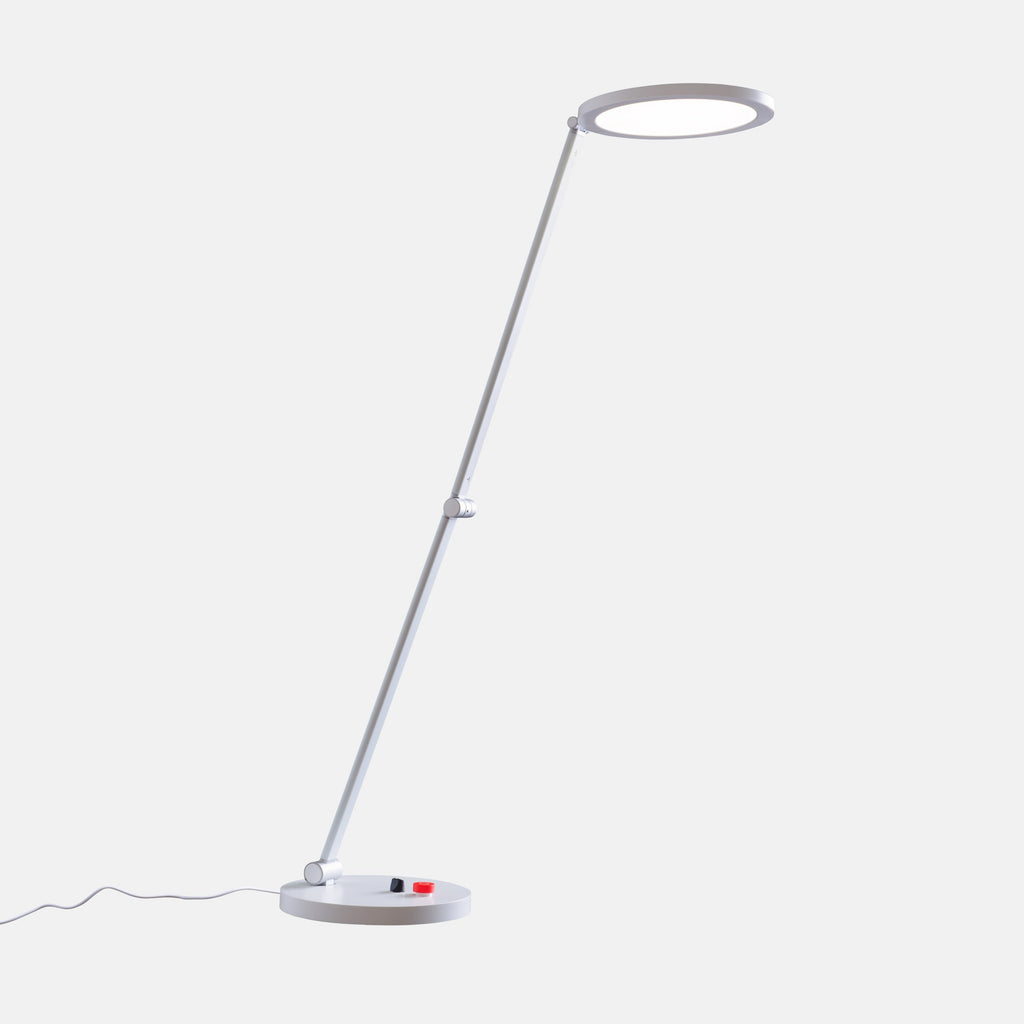 A modern adjustable lamp with a thin silver metal stand, a circular ring-shaped head, and a small red and black switch on the base, shown against a light gray background.