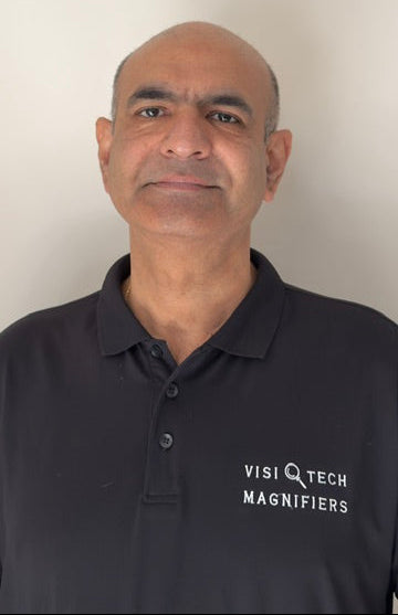 A man wearing a black polo shirt with 'VISITECH MAGNIFIERS' printed in white on the left chest, photographed against a plain off-white wall.