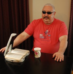 A man wearing a red graphic t-shirt and sunglasses sits behind a black table, while LyriQ is scanning a book. He looks like he is listening to audio. 