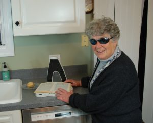 An older woman with gray hair and sunglasses stands at a kitchen counter, with a book open on LyriQ - her head is turned away so we assume she is listening. 