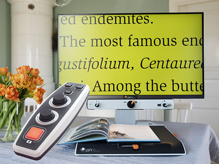 MagniLink on a table being used to read a book. Remote control is in the foreground. 