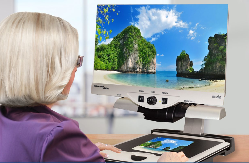 A woman with short blonde hair sits at a desk using a Merlin desktop magnifier to look at a photo displaying a tropical beach scene. The woman is wearing glasses and a purple top.