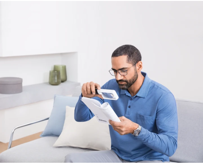A man wearing a blue polo shirt and glasses sits on a light gray sofa, using a Mobilu magnifying glass to examine a document in a bright, modern room.