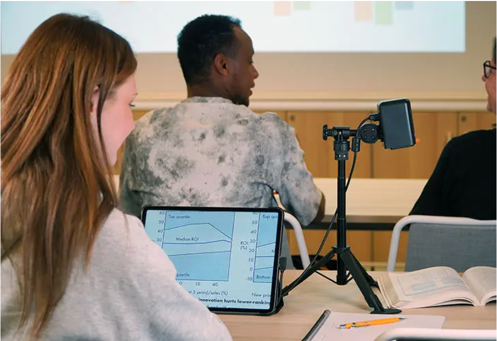 Students in a classroom setting, with one person in the foreground looking at a tablet showing a graph, another person facing a projector screen, and a MagniLink Mini on a tripod in the foreground.