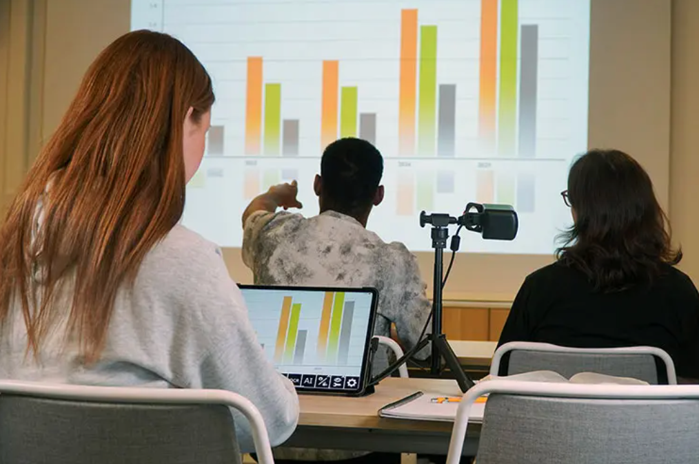 Three people seated at a table facing a large projection screen with a bar chart, with one person pointing toward the screen. A MagniLink Mini, projector, and notebook are visible on the table in a meeting or presentation setting.