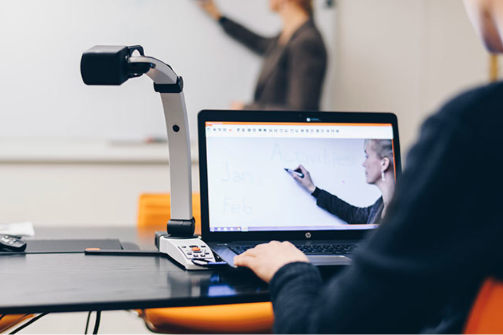 MagniLink in classroom or office setting with camera pointed forward. A person is seated at the desk using the laptop, and another person is visible and magnified in the laptop screen, using the whiteboard. 