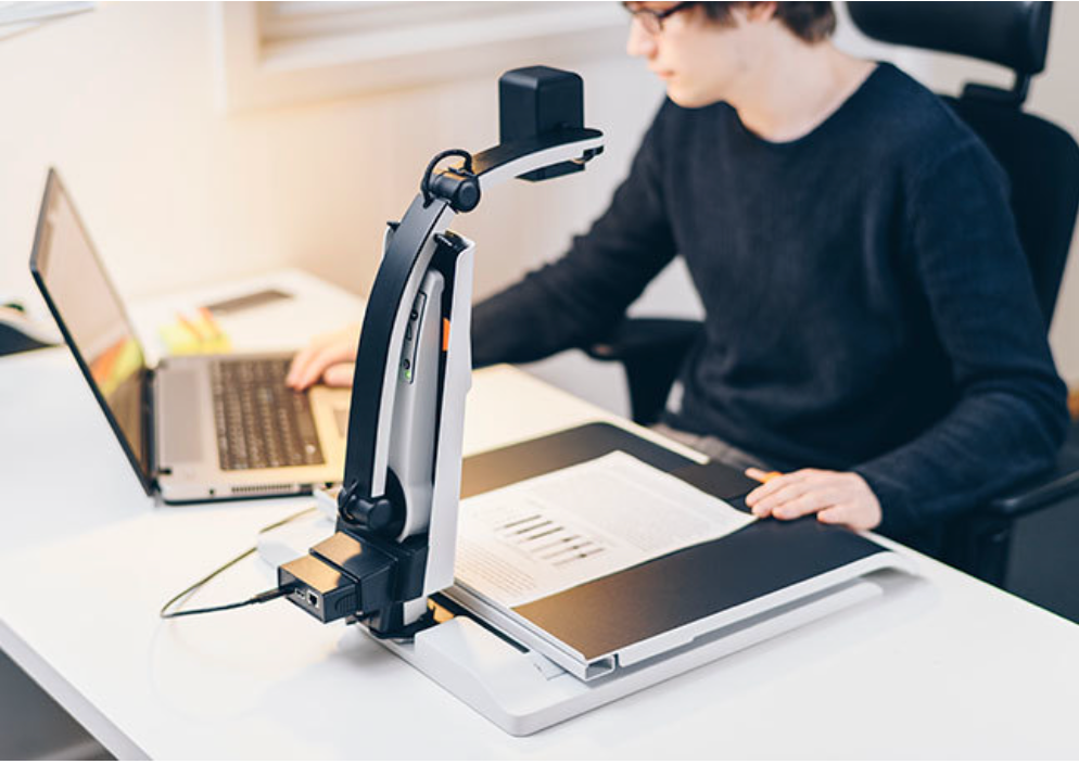 MagniLink S with camera facing the desk, magnifying a chart onto a laptop. A young man is using the laptop.