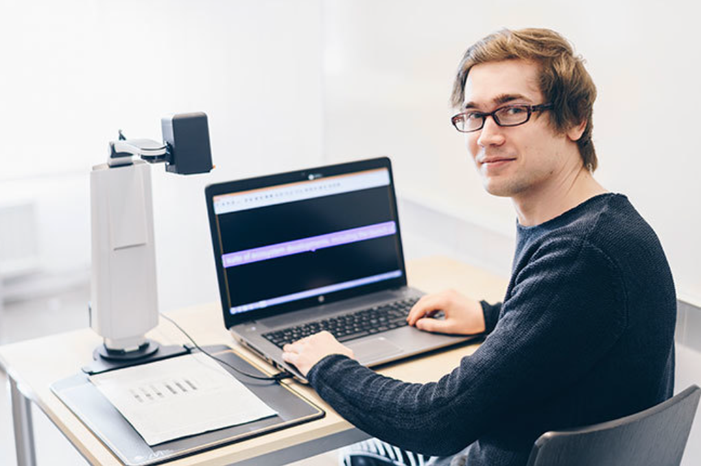 MagniLink S with camera facing the desk, magnifying a chart onto a laptop. A young man is using the laptop, and is turned to face the camera. 