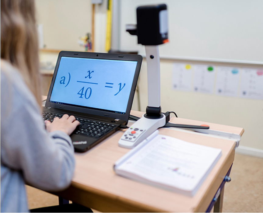 A person seated at a desk using a laptop with a math equation magnified on screen. MagniLink S is pointed down at the maths textbook in a classroom setting.