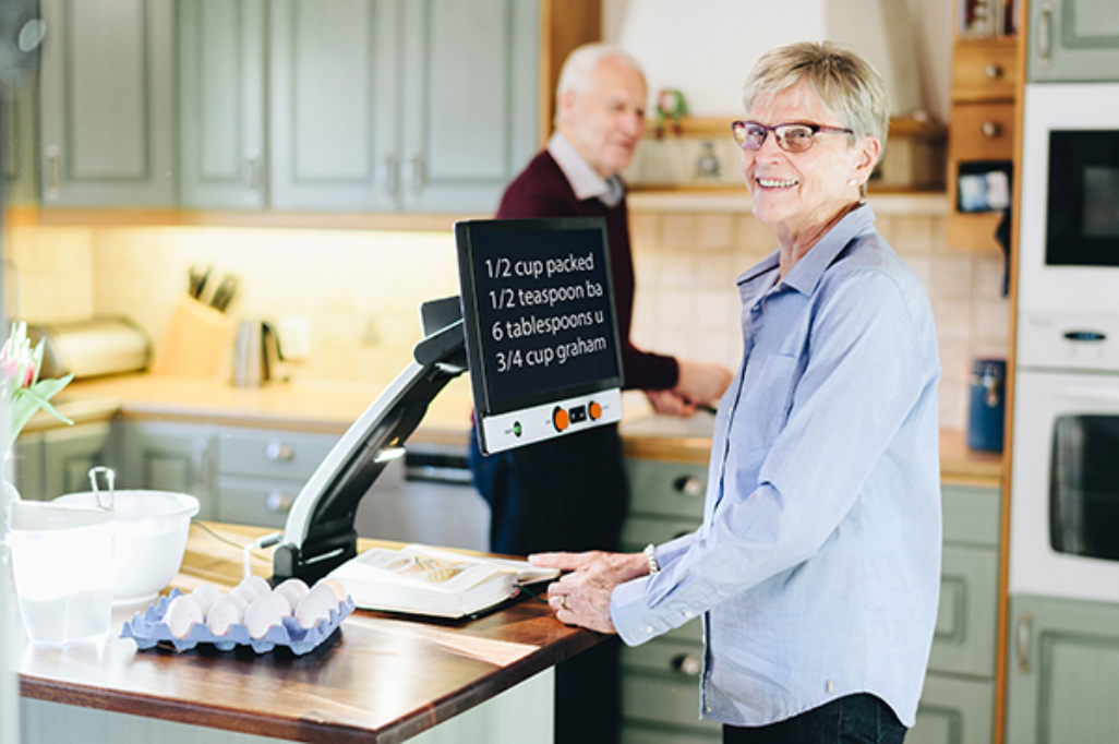 A woman in a kitchen stands at a counter using a Magnilink Vision display screen showing measurements. A man is blurred in the background of the kitchen.