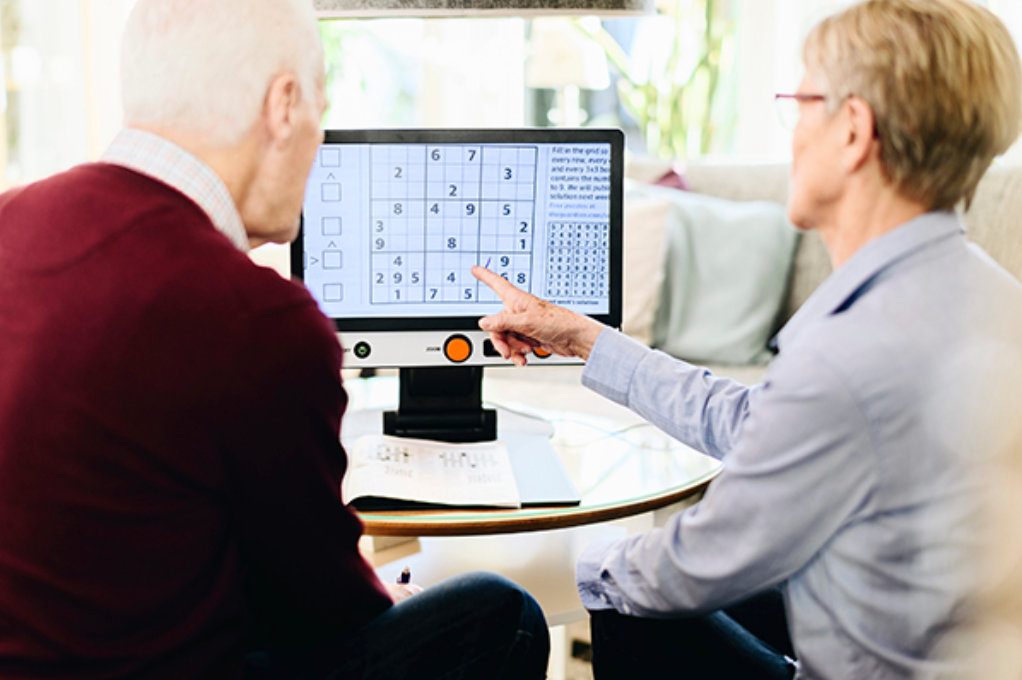 An older man and woman sit together at a table, looking at a Magnilink Vision showing a number-based puzzle game. The woman points at the screen.