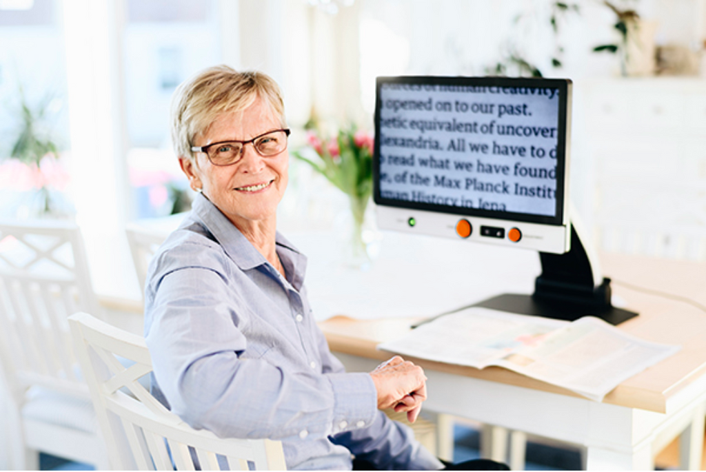 A smiling older woman with short blonde hair and glasses sits in a white chair at a desk, looking toward the camera. A Magnilink Vision displaying text is on the desk, along with papers and a bright, clean indoor background.