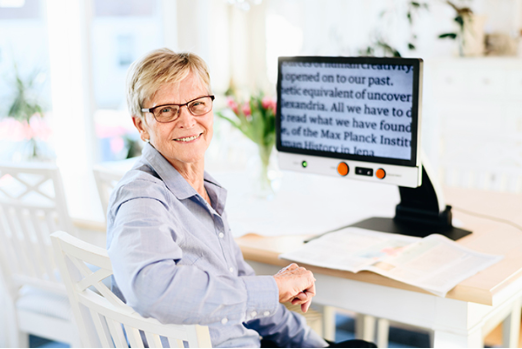A smiling older woman with short blonde hair and glasses sits in a white chair at a desk, looking toward the camera. A Magnilink Vision displaying text is on the desk, along with papers and a bright, clean indoor background.