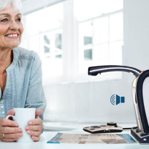 A smiling older woman sits at a table holding a white mug, with smart reader visible in a bright kitchen. Speaker symbol indicates audio playing. 