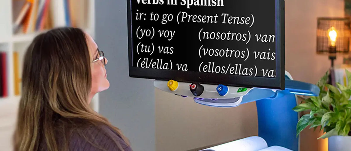 A woman wearing glasses is shown from the side, looking at magnifier text from a book placed on the desktop magnifier. The woman is in a study setting with a bookshelf and lamp in the background.