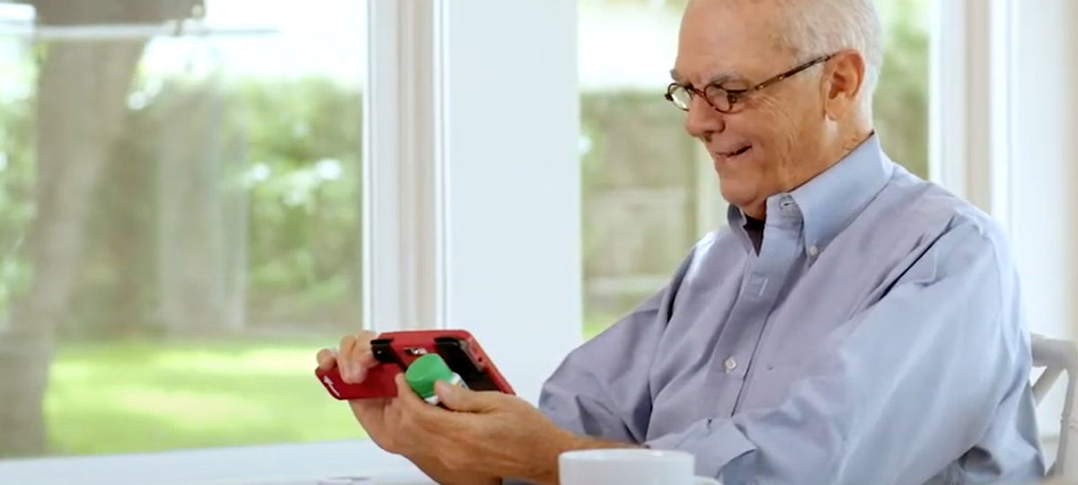 An older man with glasses and a light blue shirt sits at a table by a window, holding a red handheld magnifier, reading a medication bottle. A white mug is in the foreground, and a blurred outdoor scene is visible behind him.