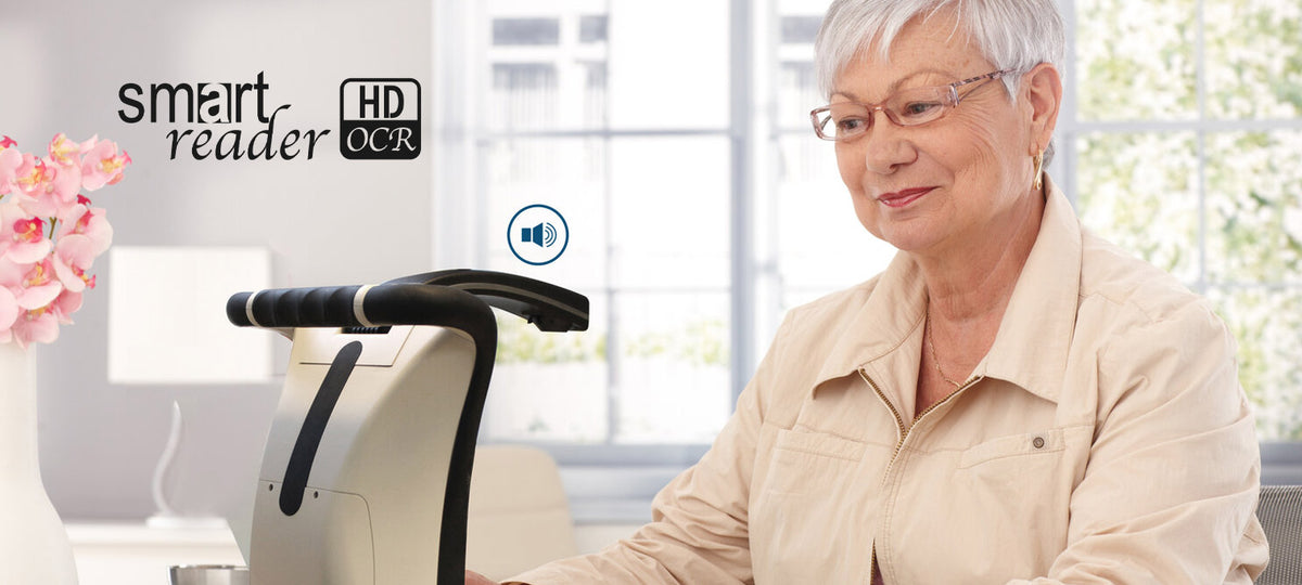 An older woman with short gray hair and glasses sits at a table using a smart reader ODR device in a bright indoor setting. The smart reader has a black handle and a light beige body. The speaker symbol is on the screen, indicating audio output. 